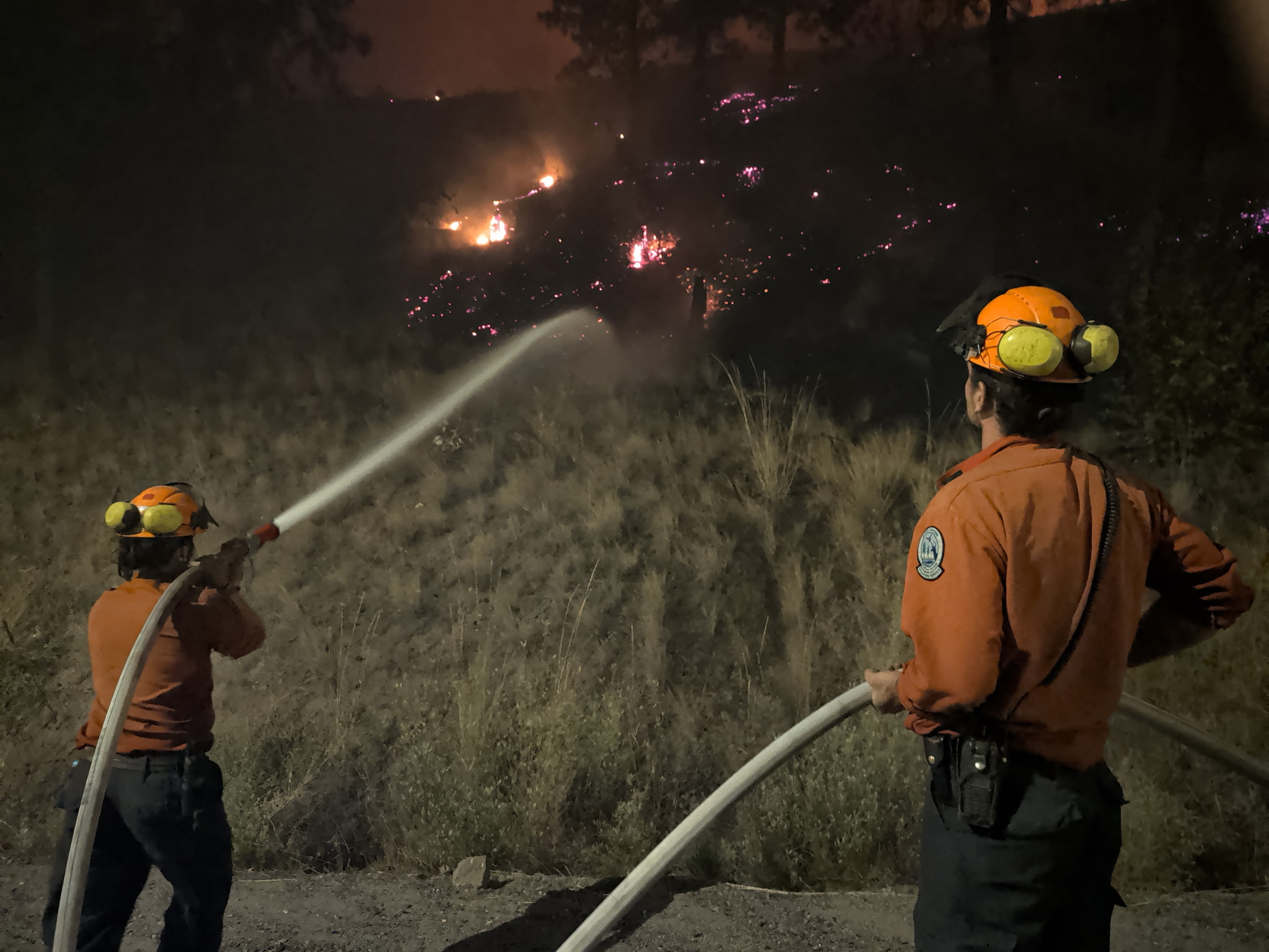 Two firefighters work a hose line at night as flames creep across a hillside.
