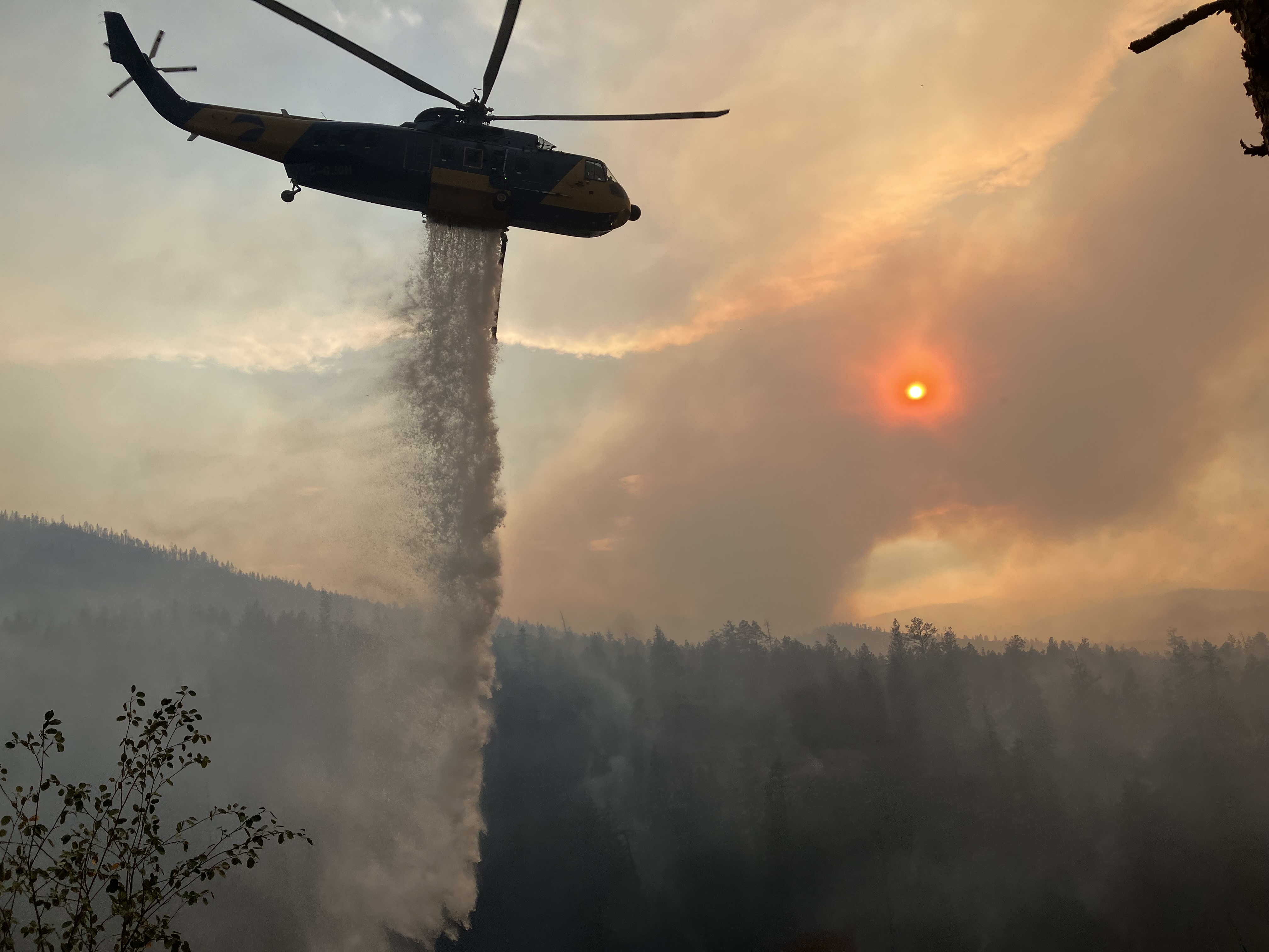 A helicopter drops water on forest below a smoke-reddened sun during the Okanagan wildfires.